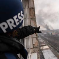 A close up photo of a blue "Press" helmet on a man pointing at a destroyed city skyline in the distance.