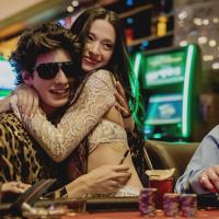 A woman passionately embraces a man sitting at a poker table inside a casino and wearing a leopard shirt and dark sunglasses.