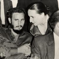 A black-and-white image of a man and a women sitting close together and talking. There is another man with glasses in the foreground but he is engaged in a separate conversation.
