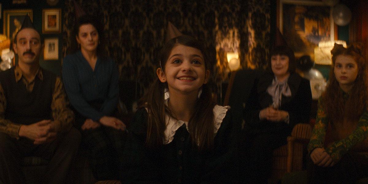 A group of five people is sitting in a dimly lit room, with the central figure being a young girl in a dark dress with a white collar. All individuals are wearing party hats, and balloons are visible in the background, indicating a celebratory occasion.