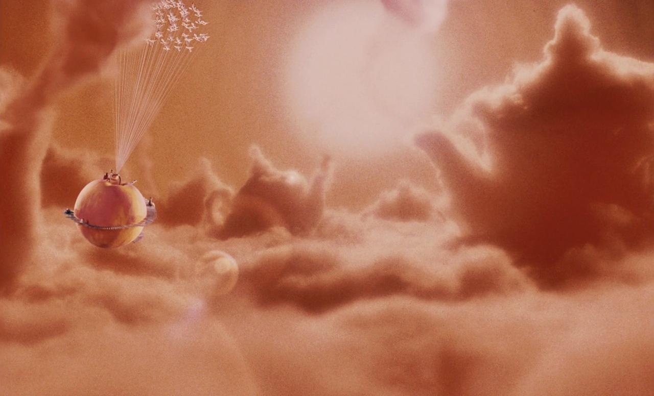 The image depicts a surreal scene with a large peach floating among clouds, lifted by multiple strings or beams extending upwards. The peach has a ring around its equator, and there are small figures or objects on top of it. Above the peach, the strings o