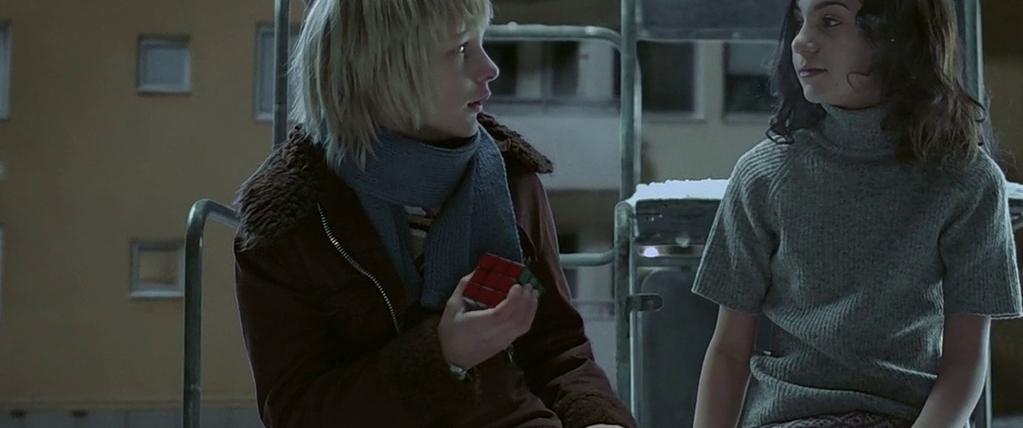 Two children are sitting next to each other. The child on the left is holding a red Rubik's Cube. Both children's faces are obscured. The background includes a building with several windows and what appears to be a metal structure, possibly a playground o