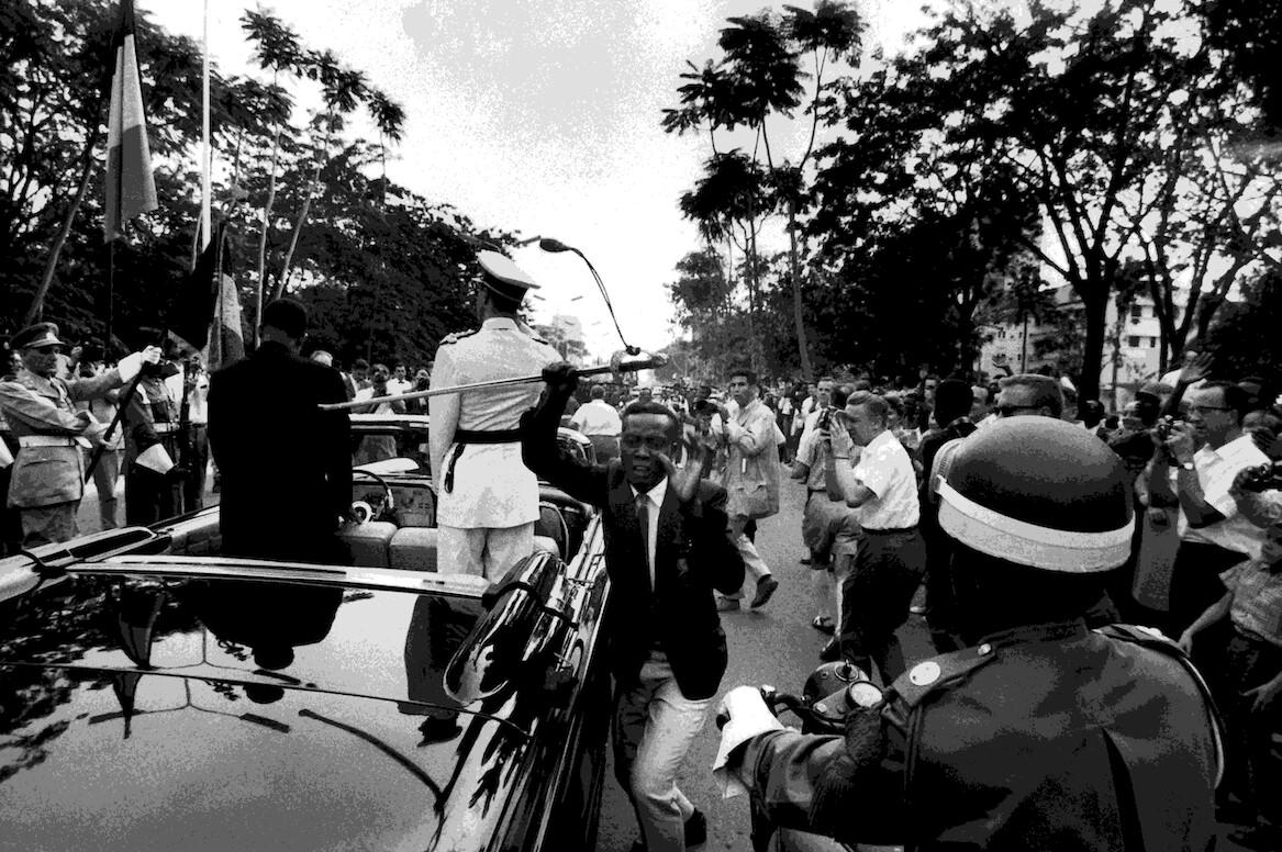 A historical scene featuring a military officer in a white uniform standing in an open-top vehicle, holding a ceremonial staff, surrounded by a crowd, with flags and trees in the background.