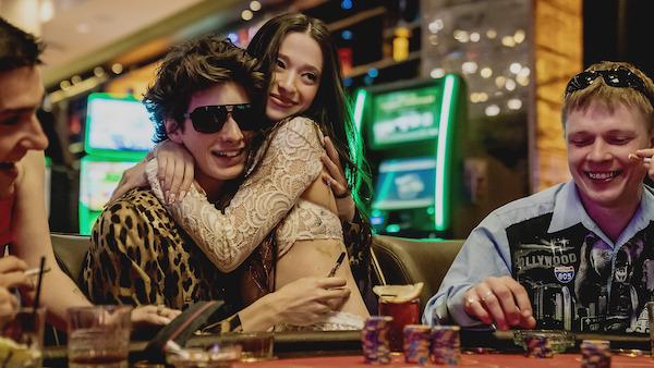 A woman passionately embraces a man sitting at a poker table inside a casino and wearing a leopard shirt and dark sunglasses.