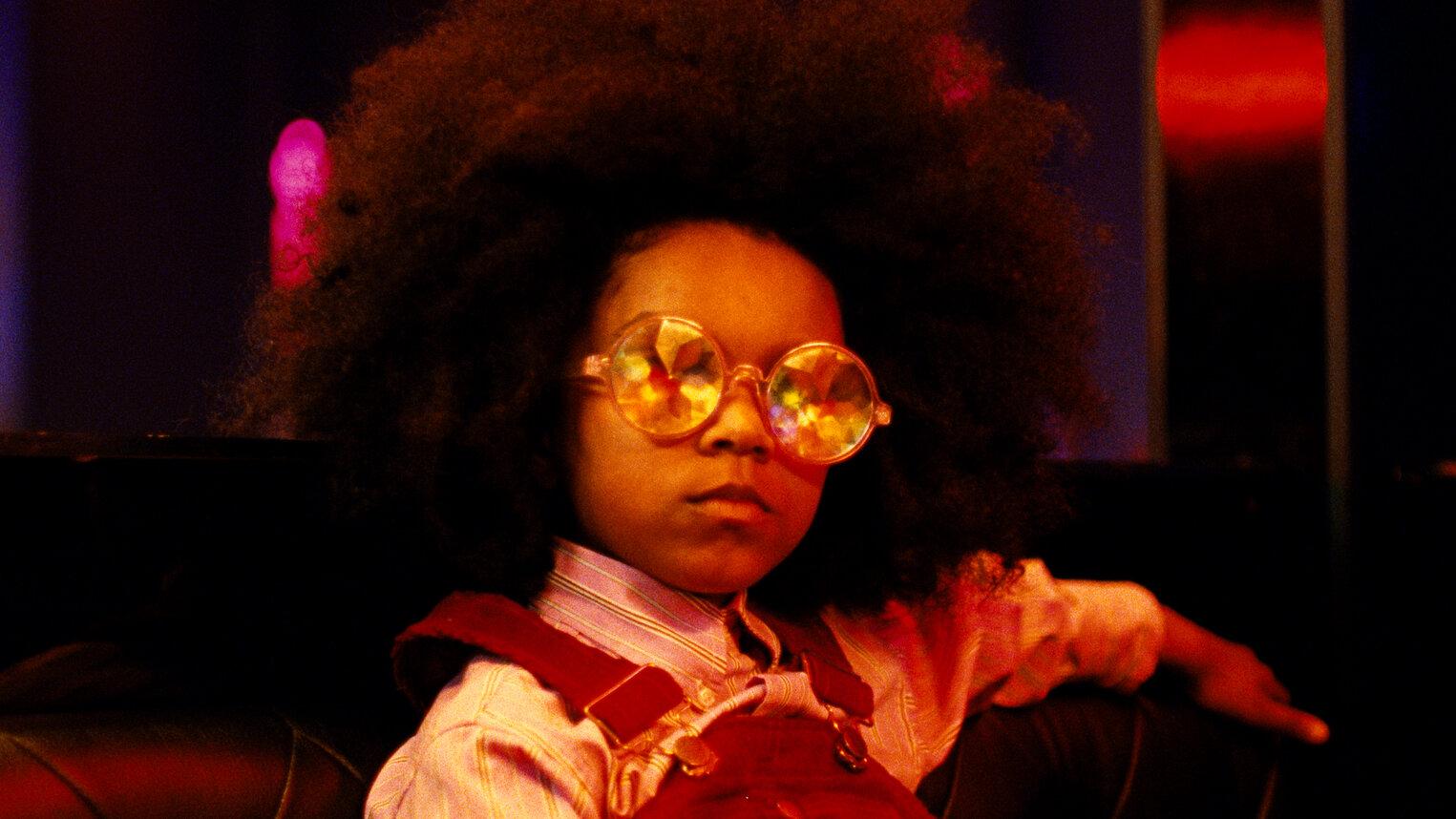A mysterious looking young girl with an afro wearing reflective round sunglasses and overalls.