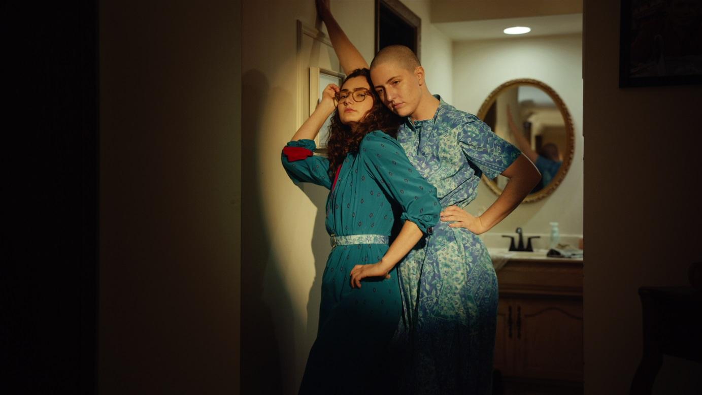 Two women -- one with a shaved head and one with long curly hair and glasses -- pose in front of a bathroom.